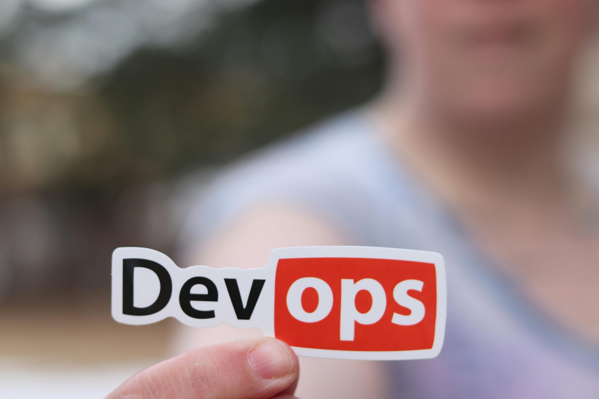 A close-up shows a person outdoors holding a DevOps sticker.